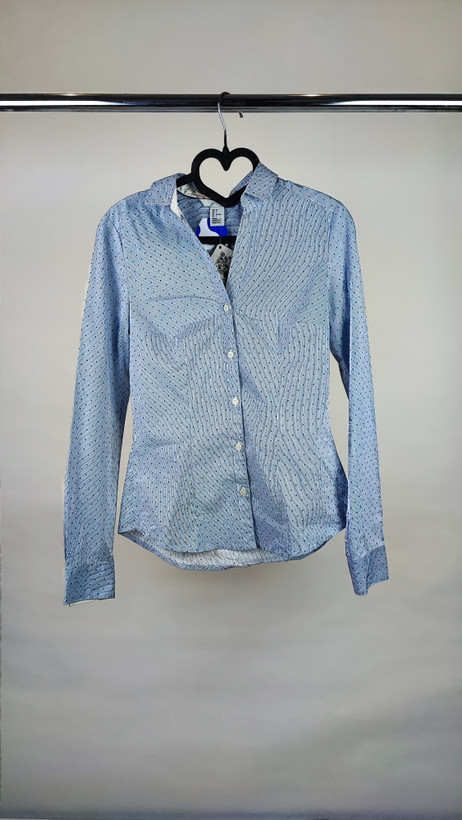 زن با شومیز آبیروشن دکمهدار و یقهبرگردان، آستینبلند و بدون طرح، در پسزمینه روشن ایستاده است