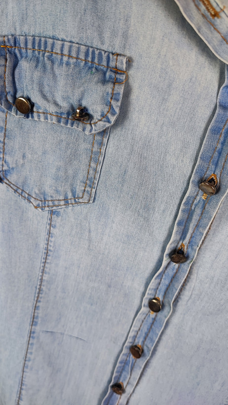 شومیز جین زنانه استوک آبی روشن، آستین بلند، یقه کلاسیک، فرم مرتب و کاربردی، مناسب استایل روزمره، ایستاده روی مدل با پسزمینه روشن.